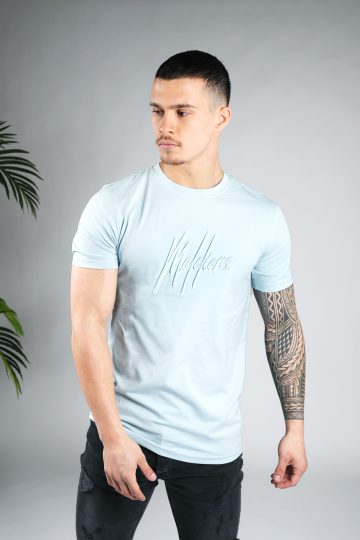 Zijaanzicht van model gekleed in lichtblauw Malelions t-shirt met het dubbel geborduurde logo in de kleuren wit en blauw op de borst. Het model heeft zijn armen langs zich.