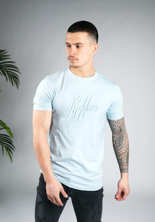 Zijaanzicht van model gekleed in lichtblauw Malelions t-shirt met het dubbel geborduurde logo in de kleuren wit en blauw op de borst. Het model heeft zijn armen langs zich.