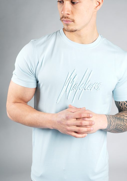 Vooraanzicht van model gekleed in lichtblauw Malelions t-shirt met het dubbel geborduurde logo in de kleuren wit en blauw op de borst. Het model heeft zijn handen voor zich.