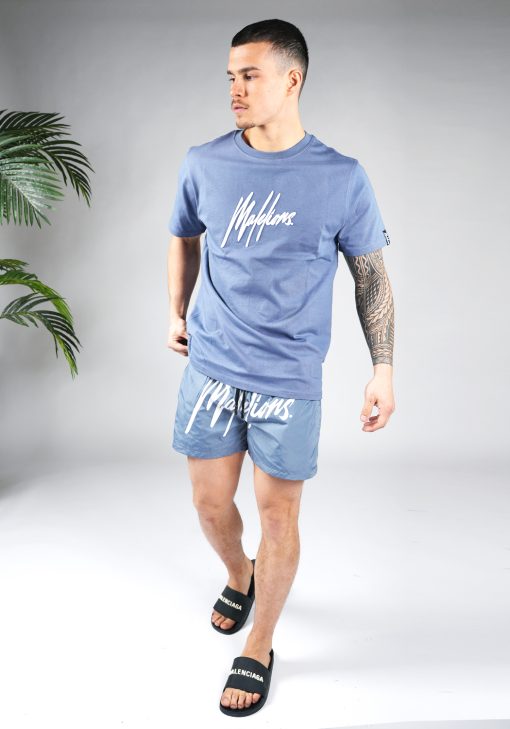 Totaal aanzicht van model gekleed in een blauw gekleurd heren T-shirt. Het T-shirt is voorzien van het MALELIONS-logo op de borst in witte kleur en een relaxed fit pasvorm. Het T-shirt wordt gecombineerd met zwarte badslippers en een blauw short.