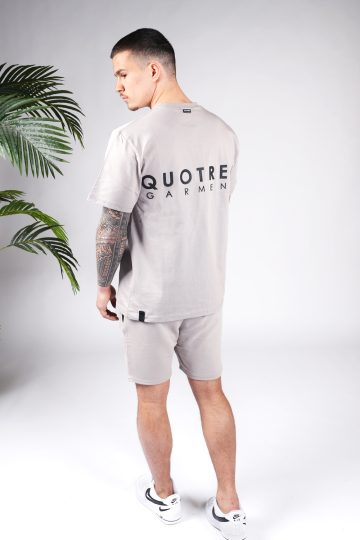Schuin achteraanzicht van model gekleed in taupe kleurige Quotrell set. De set bestaat uit jog shorts en een t-shirt. In het midden van de borst, de rug en op het linker bovenbeen staat de tekst Quotrell.