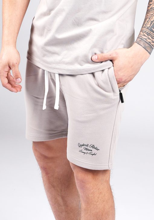 Close up broek van model gekleed in grijze Quotrell Atelier set. De set bestaat uit een t-shirt met zwarte tekst op de rug en linkerborst, en jogger shorts met tekst op de been en witte touwtjes.