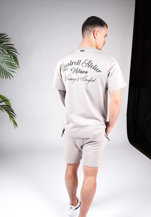Achteraanzicht van model gekleed in grijze Quotrell Atelier set. De set bestaat uit een t-shirt met zwarte tekst op de rug en linkerborst, en jogger shorts met tekst op de been en witte touwtjes.