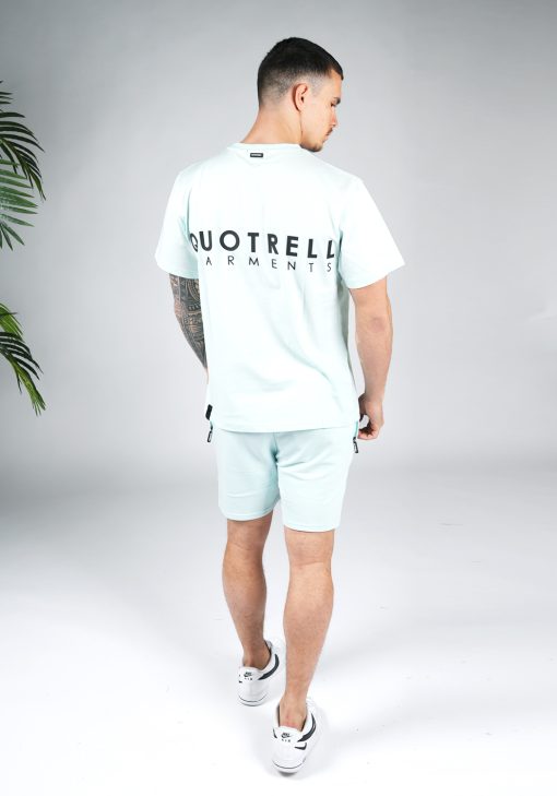 Achteraanzicht van model gekleed in mintgroene Quotrell set. De set bestaat uit een t-shirt met zwarte tekst op de rug en borst, en jogger shorts met tekst op de been.