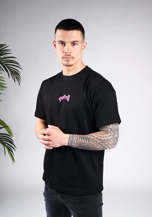 Vooraanzicht van model gekleed in een T-shirt van zwarte kleur en met een relaxed fit pasvorm. Het T-shirt is voorzien van tekst in neon roze kleur op de borst. Het model heeft zijn handen in elkaar voor het T-shirt.
