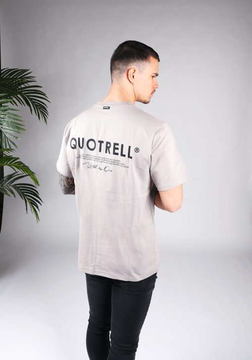 Schuin achteraanzicht van model gekleed in een heren T-shirt in taupe kleur en met ronde hals. Het T-shirt is voorzien van het QUOTRELL-logo en zwarte tekst op de rug.