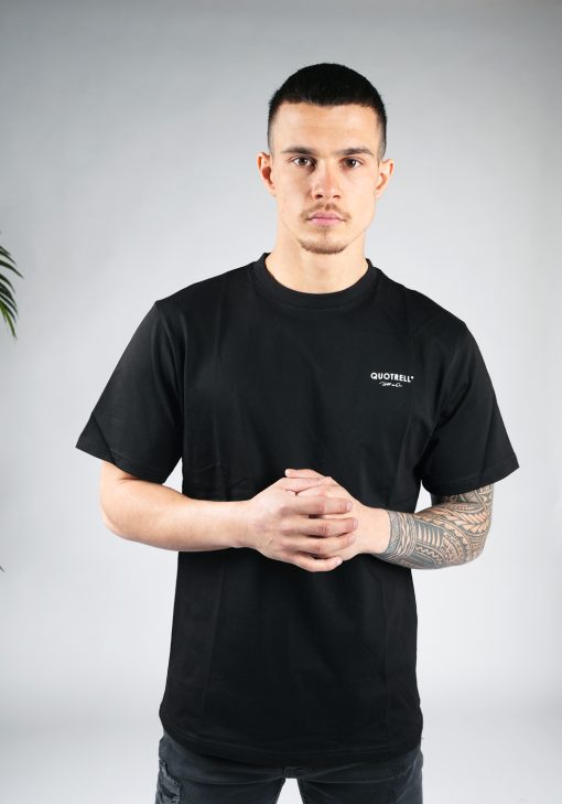 Vooraanzicht van model gekleed in een heren T-shirt in zwarte kleur en met ronde hals. Het T-shirt is voorzien van het QUOTRELL-logo en witte tekst op de linkerborst.