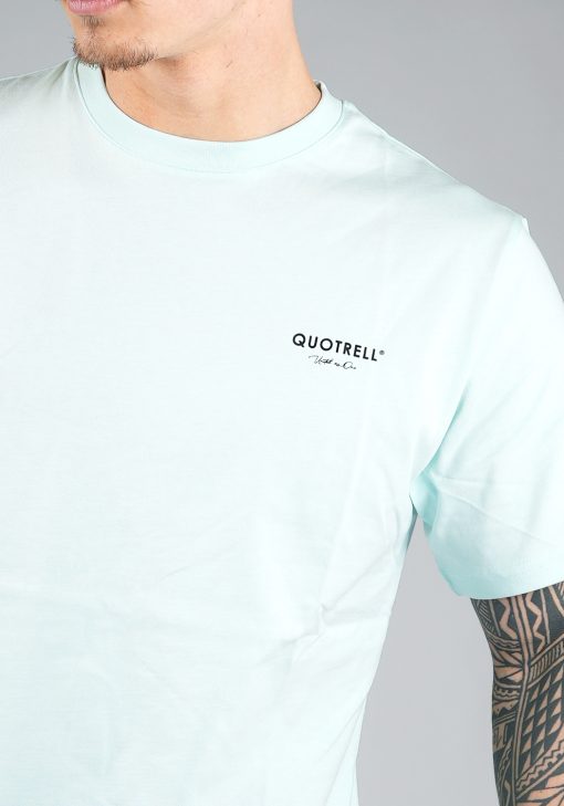 Close-up van heren T-shirt in blauwe kleur en met ronde hals. Het T-shirt is voorzien van zwarte tekst en het QUOTRELL-logo op de linkerborst.