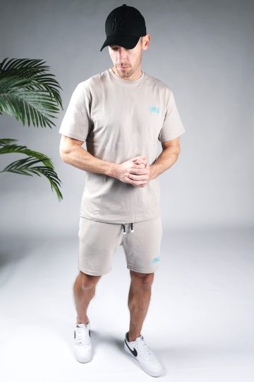 Vooraanzicht van model gekleed in grijze Quotrell set. De set bestaat uit een t-shirt met het blauwe logo op de borst en rug, en de jogger shorts met het logo op de linkerbeen.