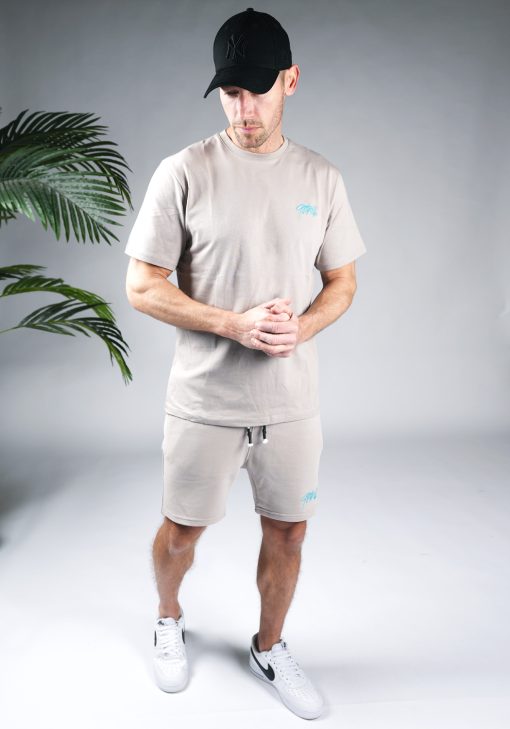 Vooraanzicht van model gekleed in grijze Quotrell set. De set bestaat uit een t-shirt met het blauwe logo op de borst en rug, en de jogger shorts met het logo op de linkerbeen.