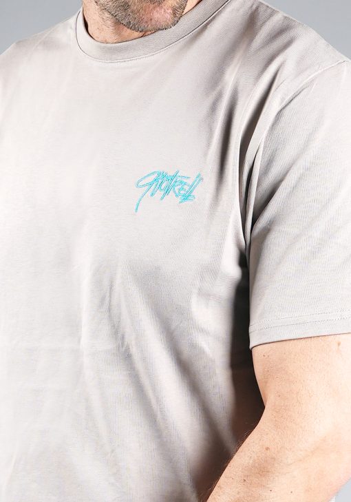 Close up shirt van model gekleed in grijze Quotrell set. De set bestaat uit een t-shirt met het blauwe logo op de borst en rug, en de jogger shorts met het logo op de linkerbeen.