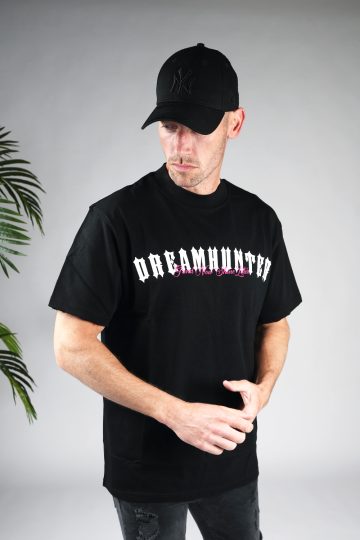 Achteraanzicht model gekleed in zwart shirt met de witte en roze tekst Dreamhunter op de borst. Het model heeft zijn handen voor zich en kijkt schuin naar de grond.