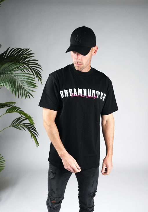 Vooraanzicht model gekleed in zwart shirt met de witte en roze tekst Dreamhunter op de borst. Het model draagt ook zwarte jeans en een zwarte pet.