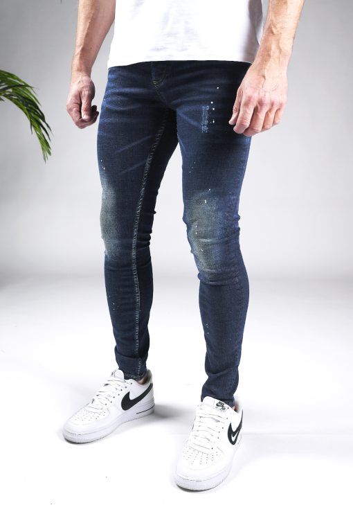 Linker zijaanzicht donkerblauwe heren skinny jeans met verfspetters en damaged afwerken, gemaakt van stretch stof. Voorzien van vier zakken, knoopsluiting en riemlussen. Gecombineerd met wit T-shirt en witte sneakers.