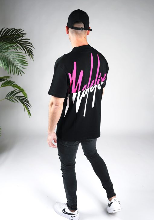 Achteraanzicht model gekleed in zwart shirt met het split Malelions logo op de rug. De bovenste helft van het logo is roze en de onderste helft is wit. Het model draagt ook zwarte jeans en een zwarte pet.