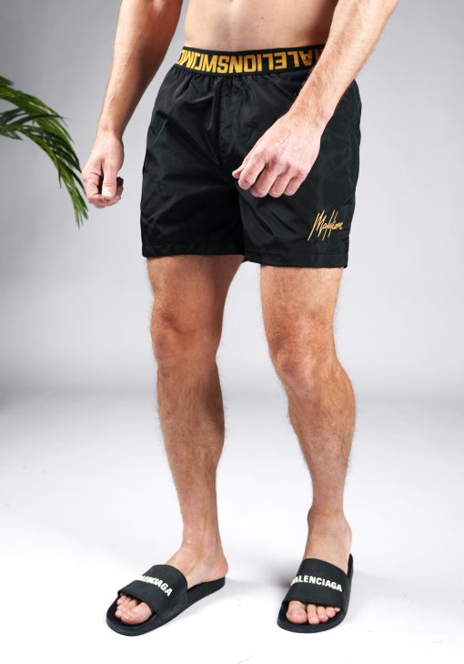 Schuin vooraanzicht onderlichaam van model gekleed in zwarte Malelions zwembroek met gouden details bij de tailleband en het gouden logo op de been.